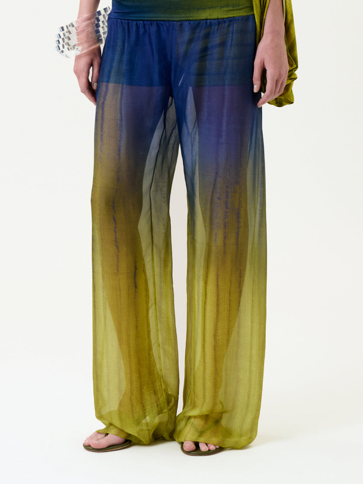 Wearing the Lene Pant—sheer, loose pants featuring a blue-to-yellow gradient—paired with flip-flops and a partially visible green top. A sheer green fabric is draped over one arm, accessorized with a chunky clear and blue bracelet on the wrist.