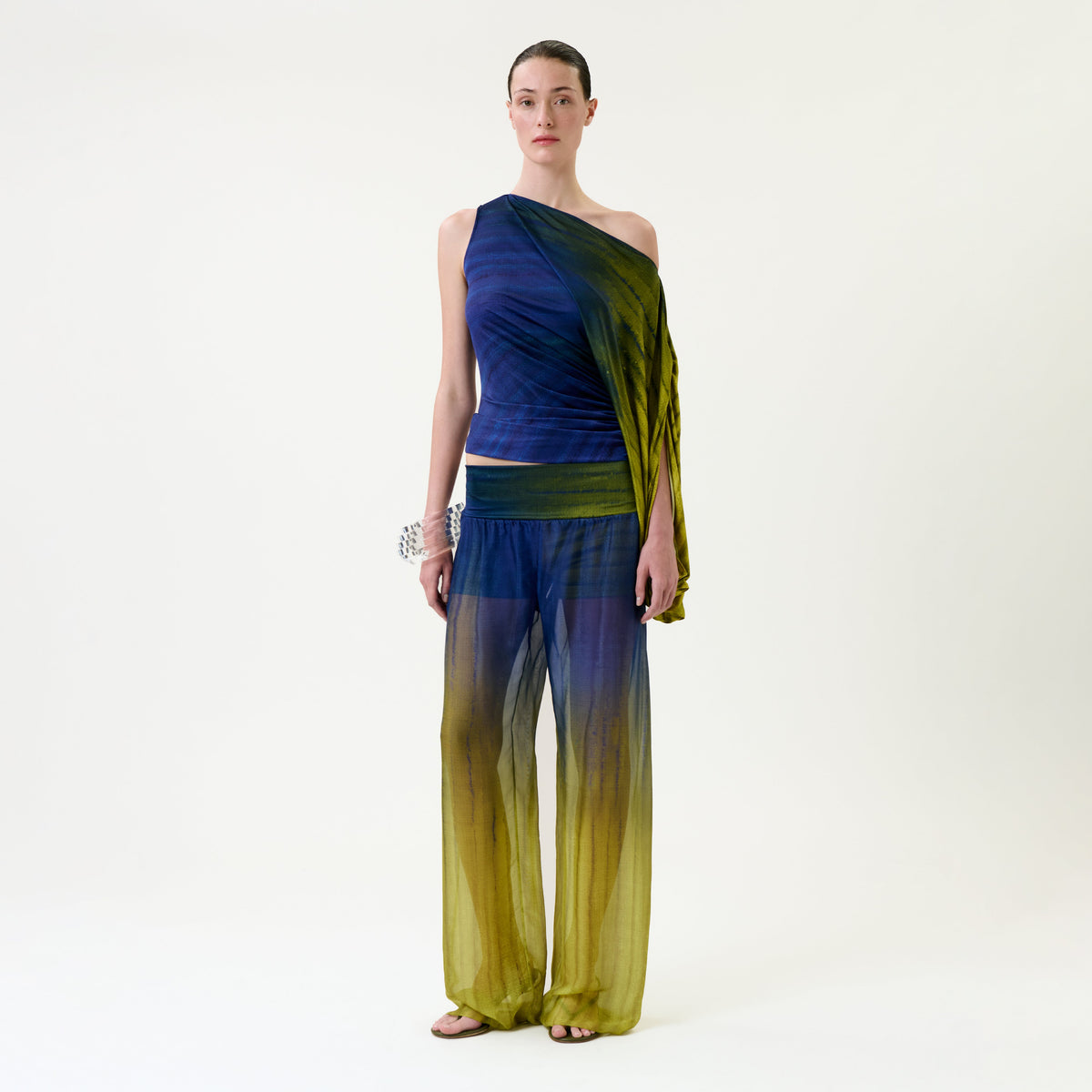 Wearing the Lene Pant in a blue and yellow gradient, paired with a sheer one-shoulder top, the individual stands against a plain light background. Their hair is pulled back and they accessorize with a chunky bracelet.