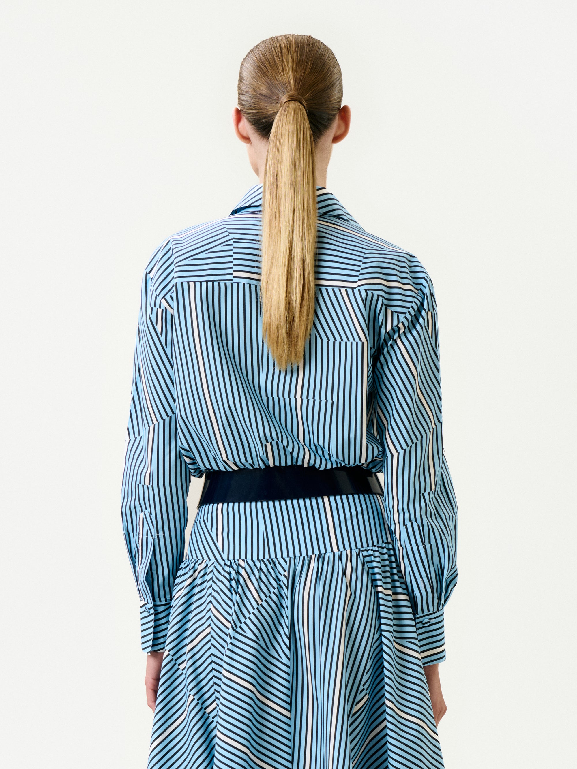 A person with long, straight blonde hair in a ponytail is seen from behind wearing the Melva Blouse—blue and white vertical stripes, crafted from 100% cotton Italian fabric—against a plain white background.