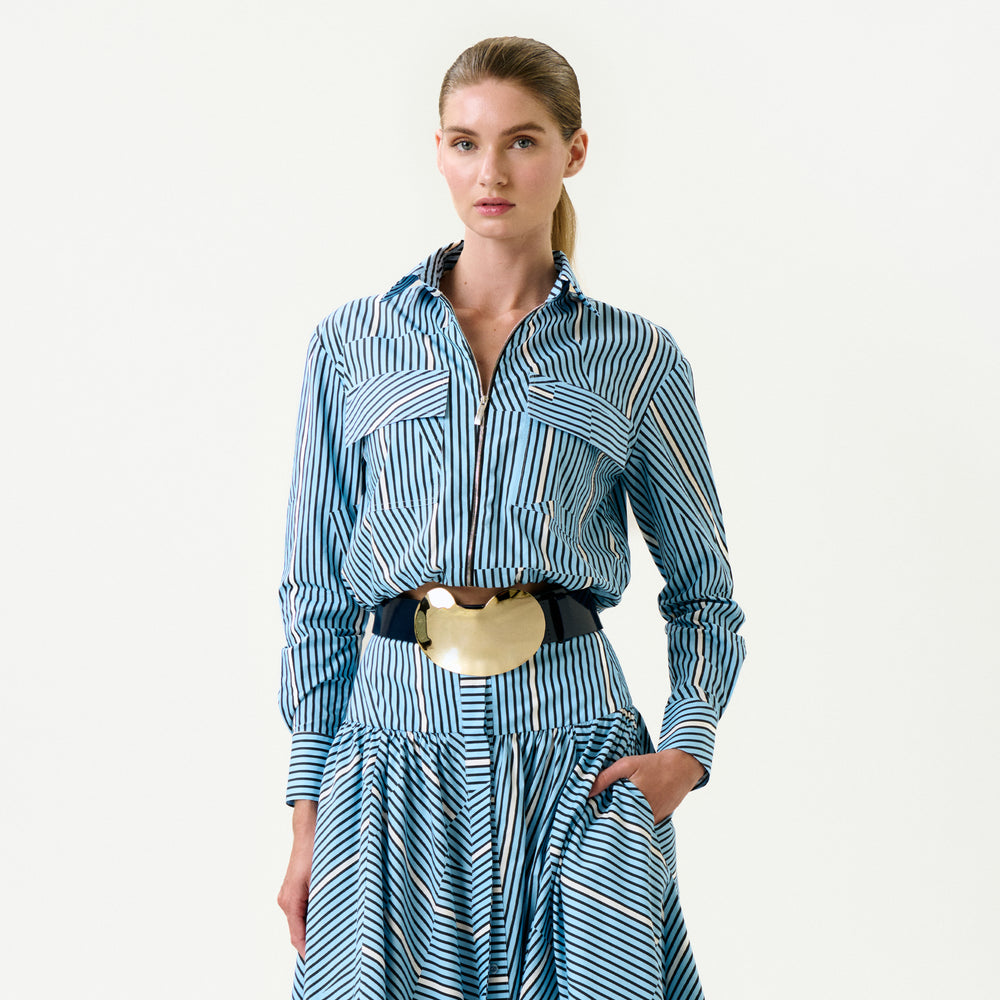 A woman poses against a plain background wearing the Melva Blouse—blue and white stripes, Italian fabric, long sleeves, a collared neckline, wide gold belt. Her hair is in a low ponytail and one hand rests in her skirt pocket.