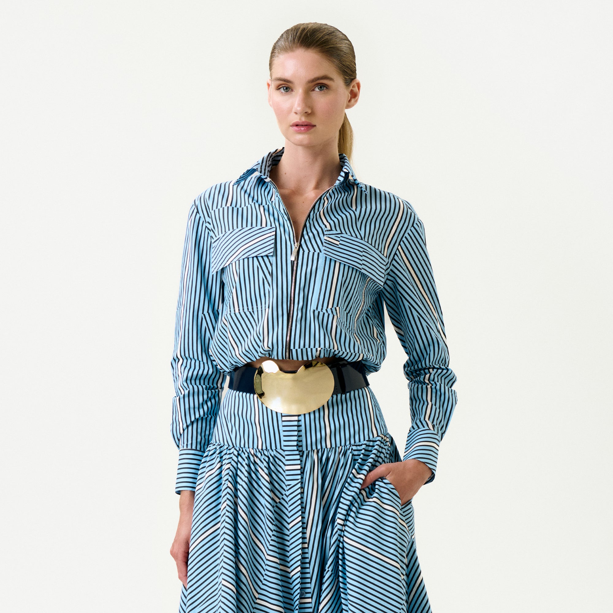 A woman poses against a plain background wearing the Melva Blouse—blue and white stripes, Italian fabric, long sleeves, a collared neckline, wide gold belt. Her hair is in a low ponytail and one hand rests in her skirt pocket.