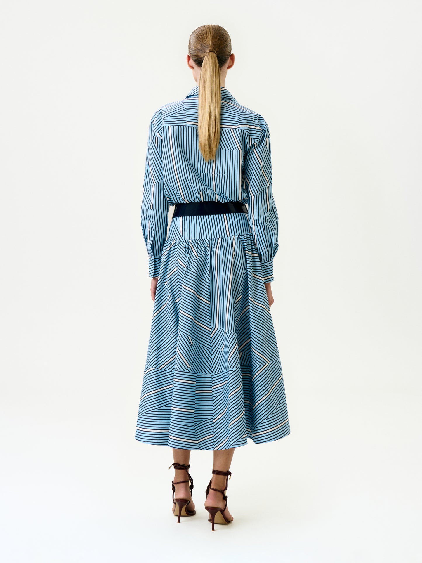 A person with long blond hair in a low ponytail wears the Milva Skirt, styled with a black belt and brown high-heeled sandals, facing away against a plain white background.