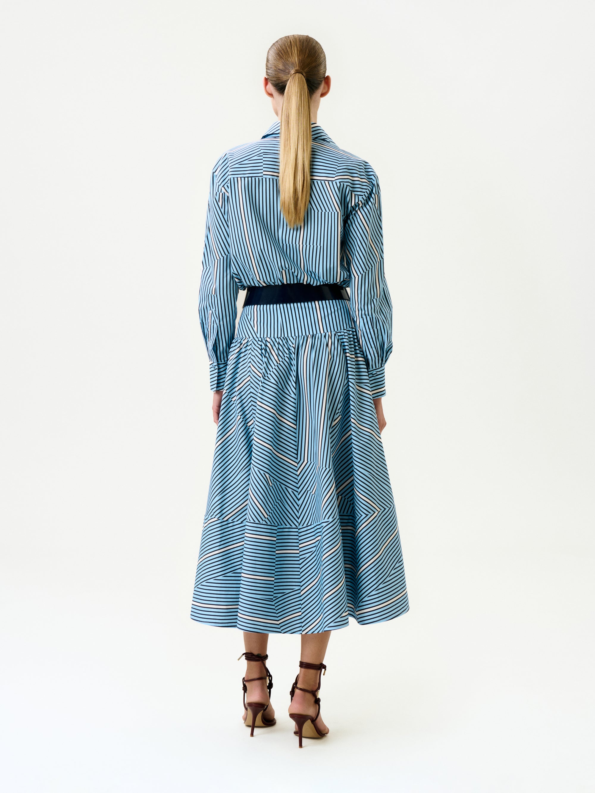 A person with long blond hair in a low ponytail wears the Milva Skirt, styled with a black belt and brown high-heeled sandals, facing away against a plain white background.