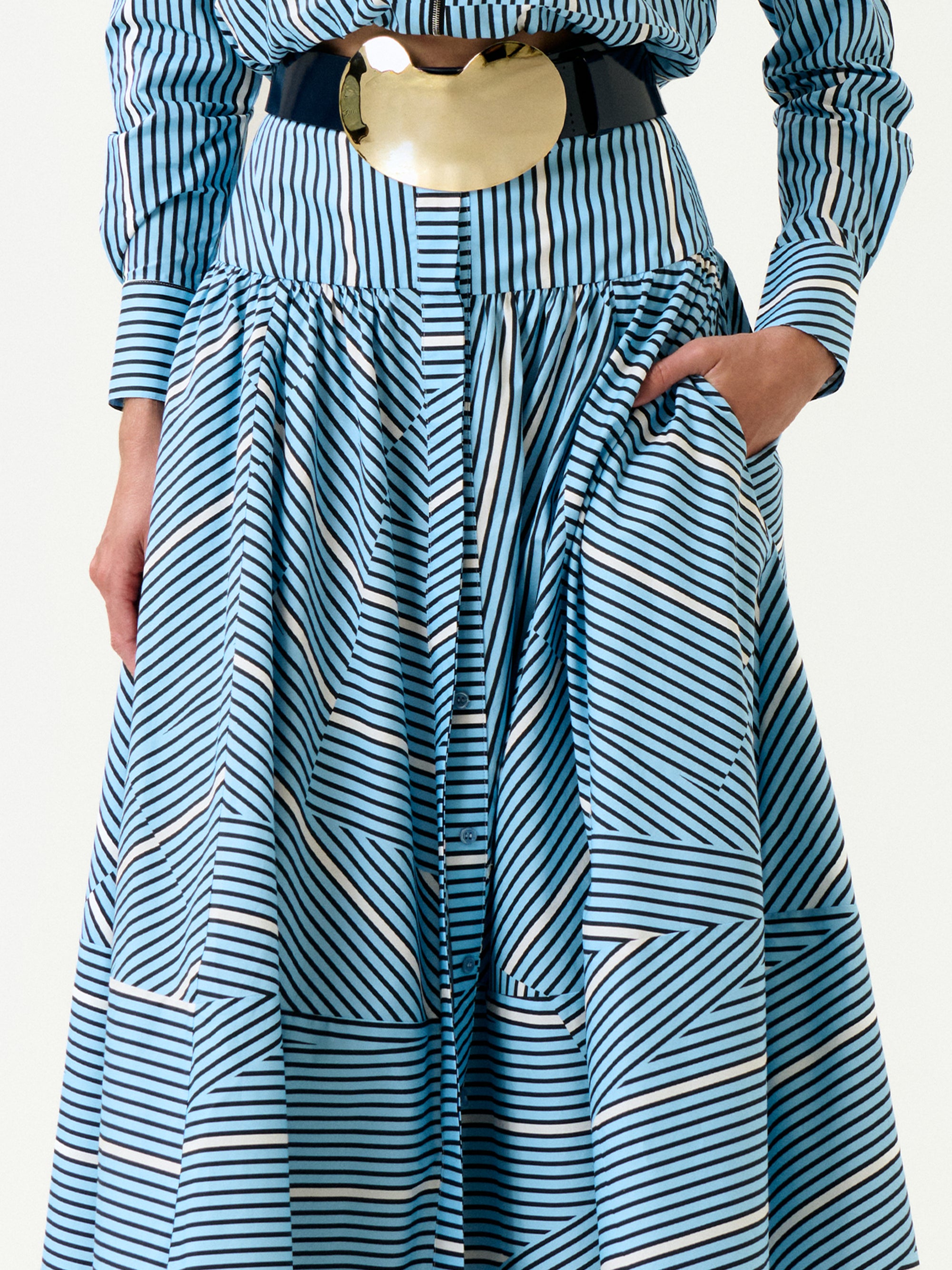 Wearing the Milva Skirt, a blue and white striped piece with geometric patterns made from Italian fabric, cinched at the waist with a wide black belt and gold buckle, one hand in the pocket.