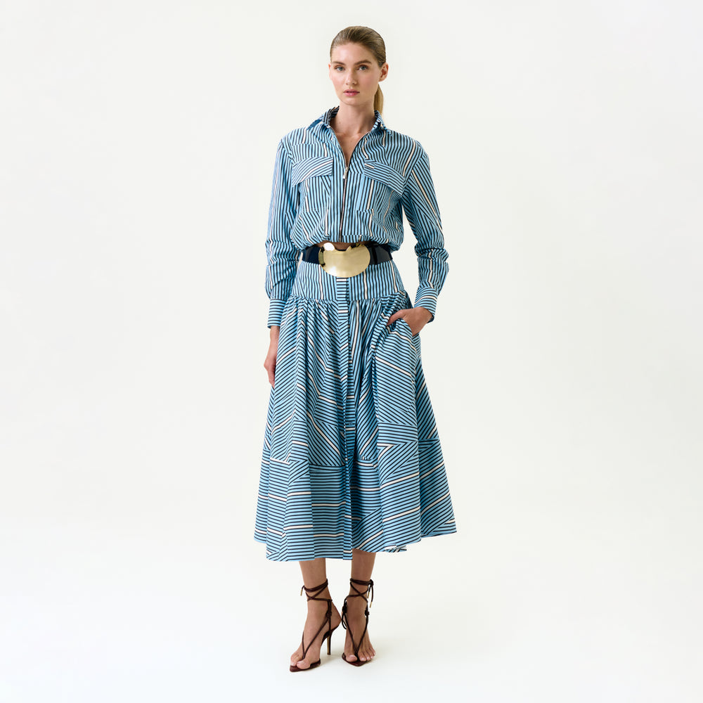 A woman stands against a plain background wearing the Milva Skirt—blue and white striped, crafted from 100% cotton Italian fabric—paired with a coordinating blouse, metallic belt, and strappy high-heeled sandals.