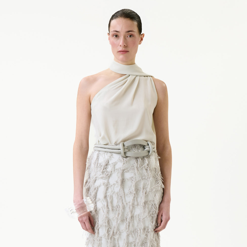 Wearing the Onora Blouse, an off-white, asymmetrical sleeveless top, and a textured skirt with a belt, a person stands against a plain light background, looking directly ahead.