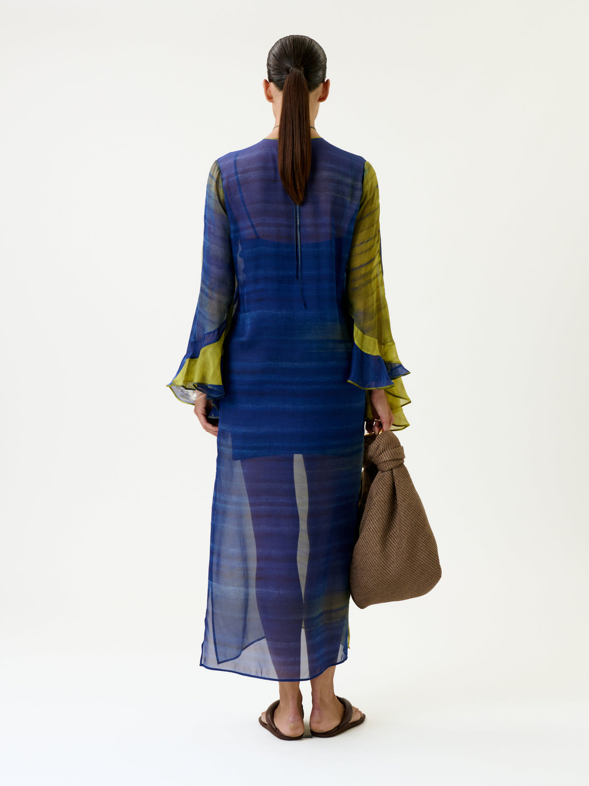 A woman with a low ponytail stands facing away against a plain white background, wearing the Aries Dress—a sheer blue and green long-sleeve dress—paired with brown flip-flops and holding a textured brown bag.