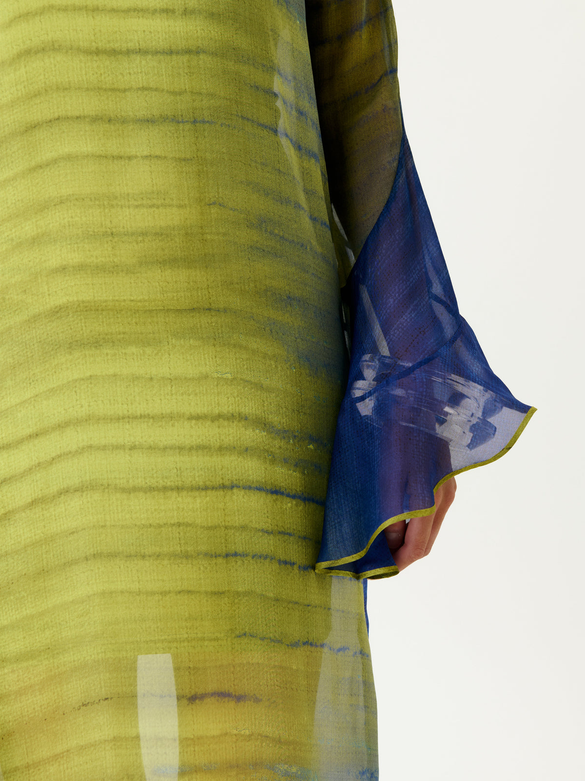 A close-up of a person wearing the Aries Dress, featuring a sheer yellow-green fabric with horizontal stripes and a sheer blue sleeve, hand partially visible.