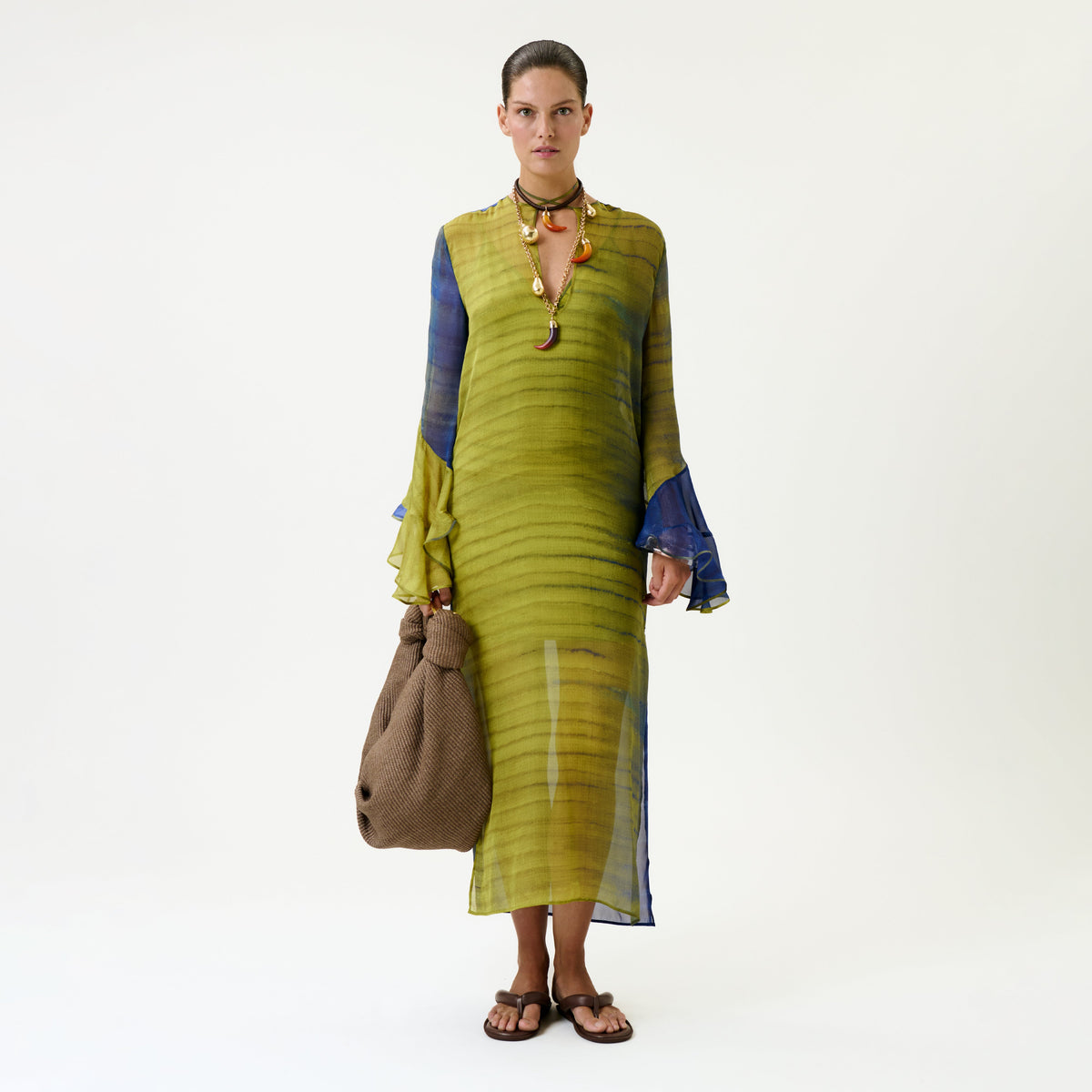A person wearing the Aries Dress, a sheer green and blue long-sleeve dress, along with brown sandals, a necklace, and holding a large brown textured bag, stands against a plain white background.