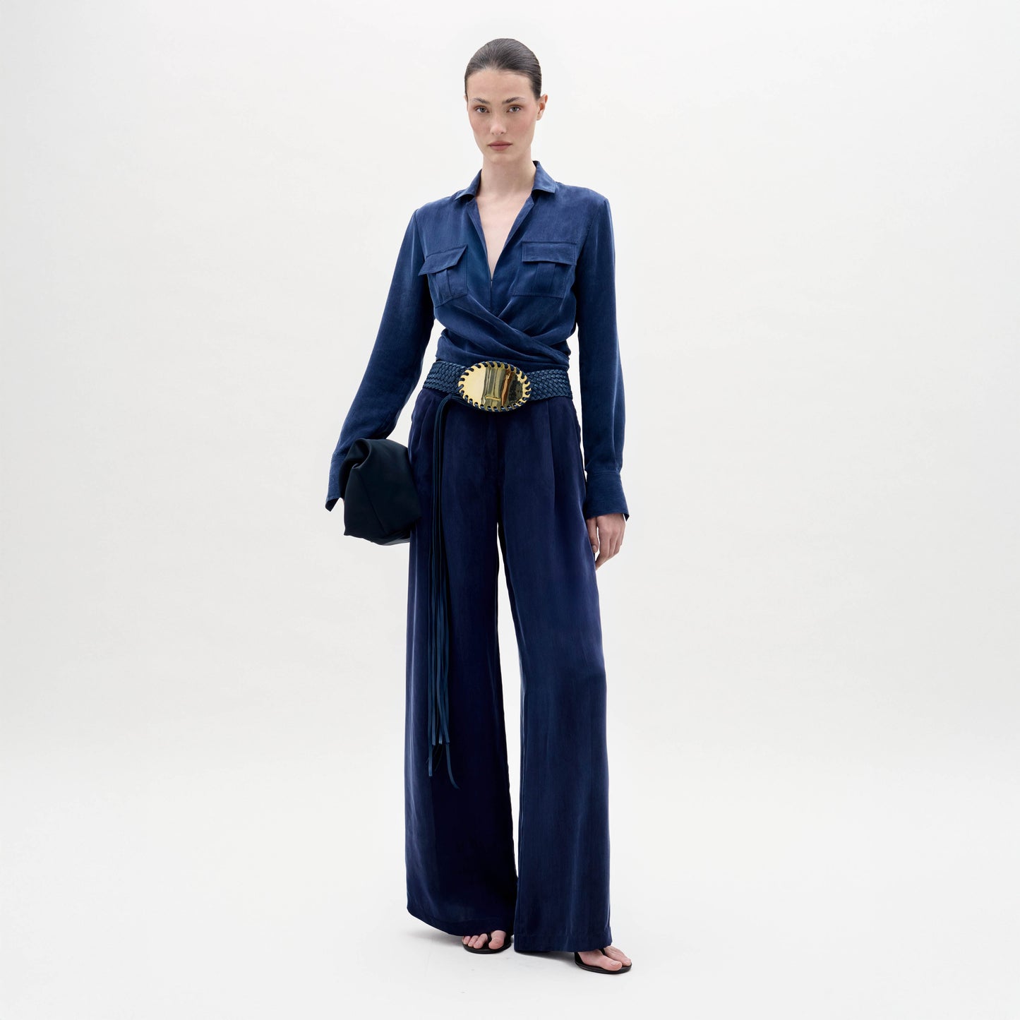 A woman stands against a plain background wearing the Opal Pant, paired with a deep blue long-sleeve blouse, a large gold-detailed belt, sandals, and holding a small clutch.