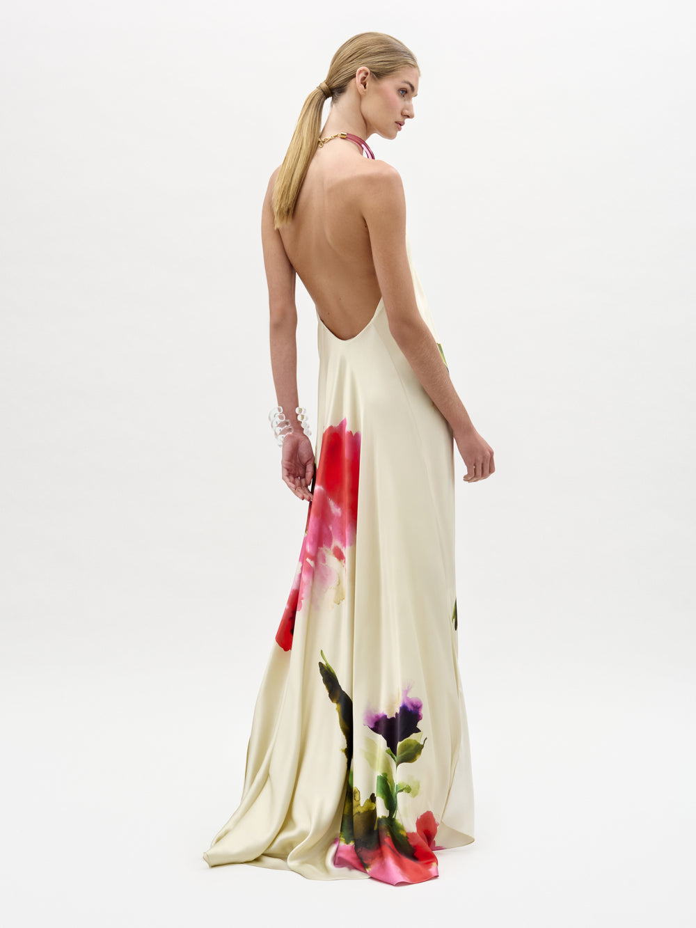 A woman with long, straight blonde hair in a low ponytail wears the cream-colored Jesse Dress—a backless maxi with a halter neckline and flowing train adorned with bold, watercolor floral prints—standing against a white background.
