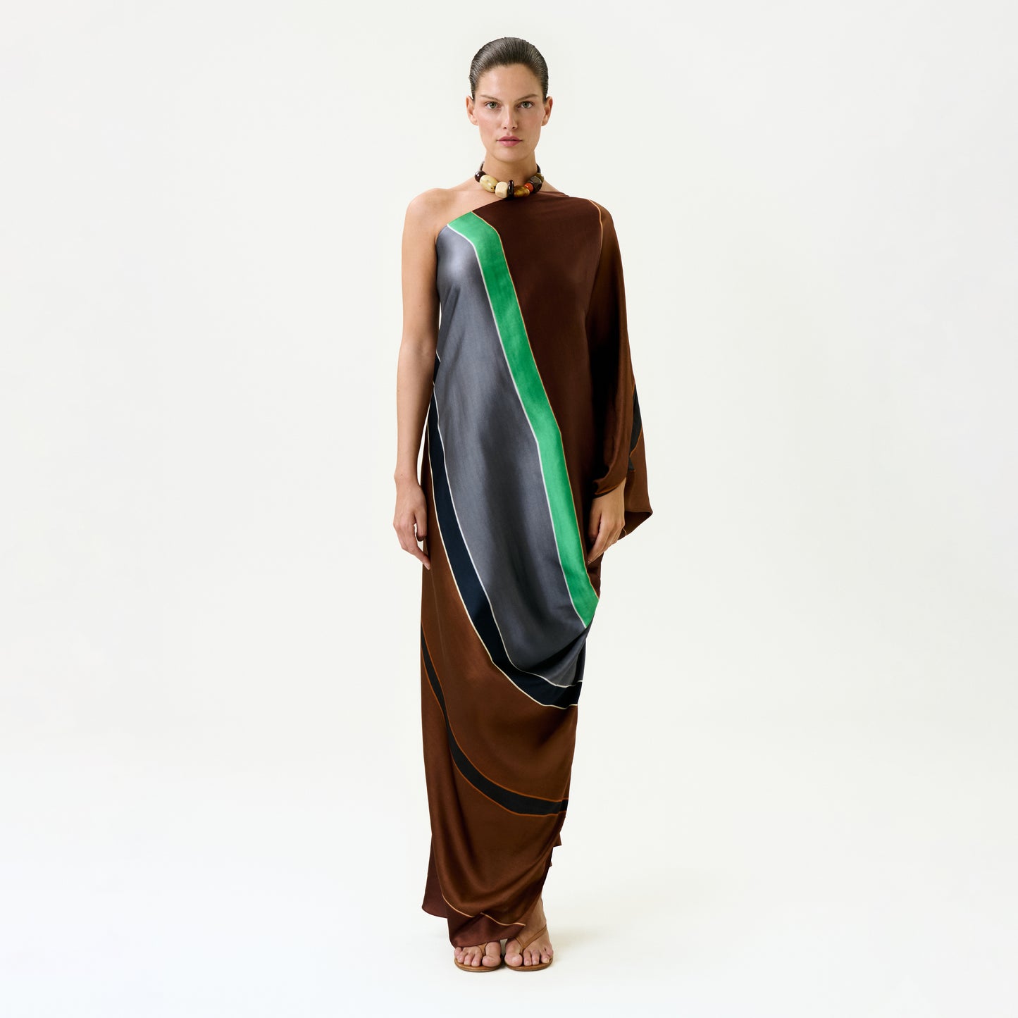 A woman stands against a plain white background wearing the Inu Tunic, a one-shoulder maxi dress with green, gray, and black diagonal stripes in luxurious Italian fabric. She is barefoot, hair pulled back, and wears a necklace.