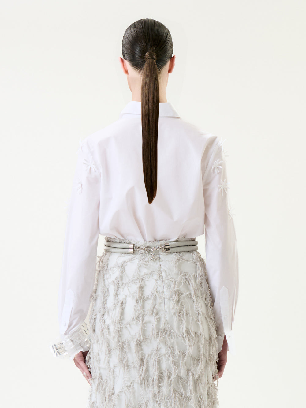 A person with long, straight brown hair in a ponytail is seen from behind against a plain background, wearing the Naroa Shirt—a white top featuring textured patterns—paired with a high-waisted, feathered white skirt.