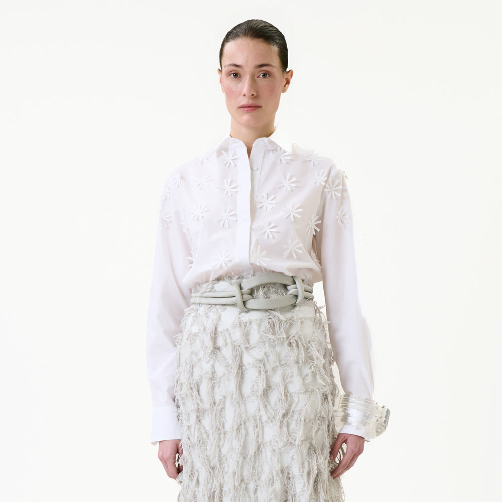 A woman wearing the Naroa Shirt, featuring floral patterns, paired with a textured, fringed white skirt stands against a plain white background, looking directly at the camera.