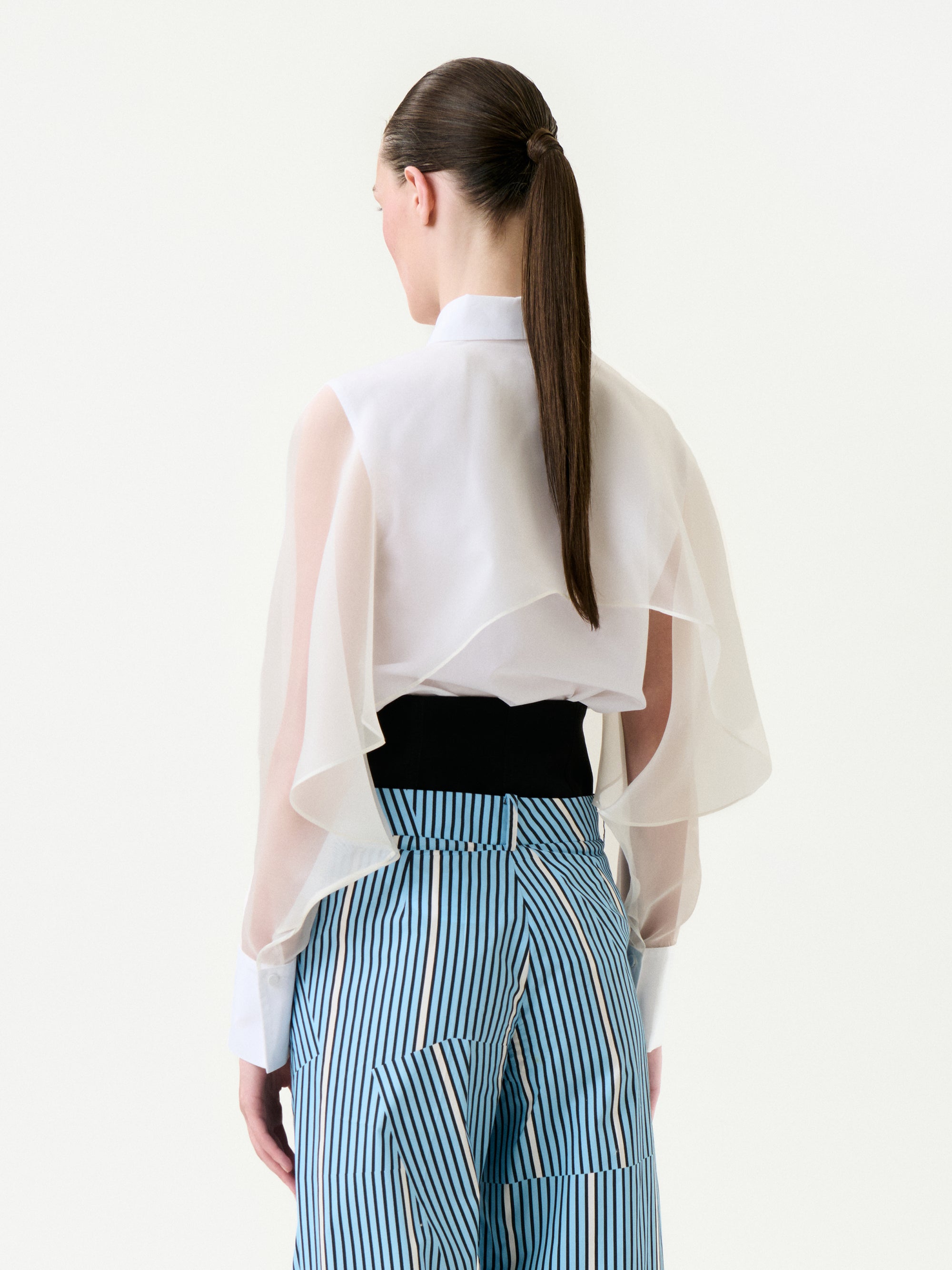 A person faces away, wearing the Nira Shirt—a sheer white blouse with flowing sleeves—tucked into high-waisted blue and white striped cotton pants. Their long brown hair is styled in a sleek ponytail.