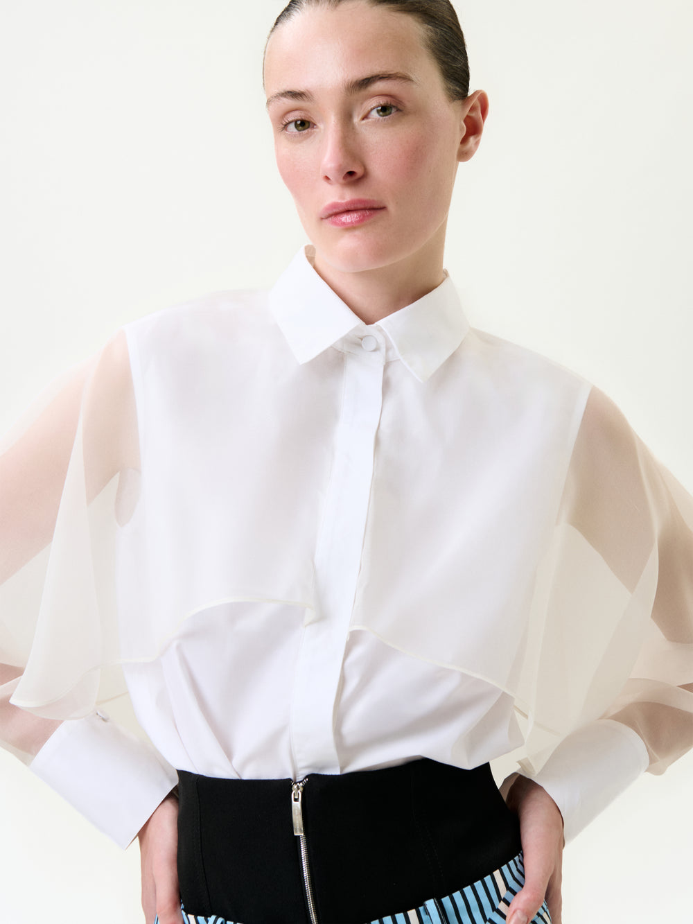 A woman with slicked-back hair wears the Nira Shirt—a sheer, layered-sleeve blouse made from 100% cotton Portuguese fabric—and a high-waisted black skirt with blue and white stripes, standing before a plain light background.