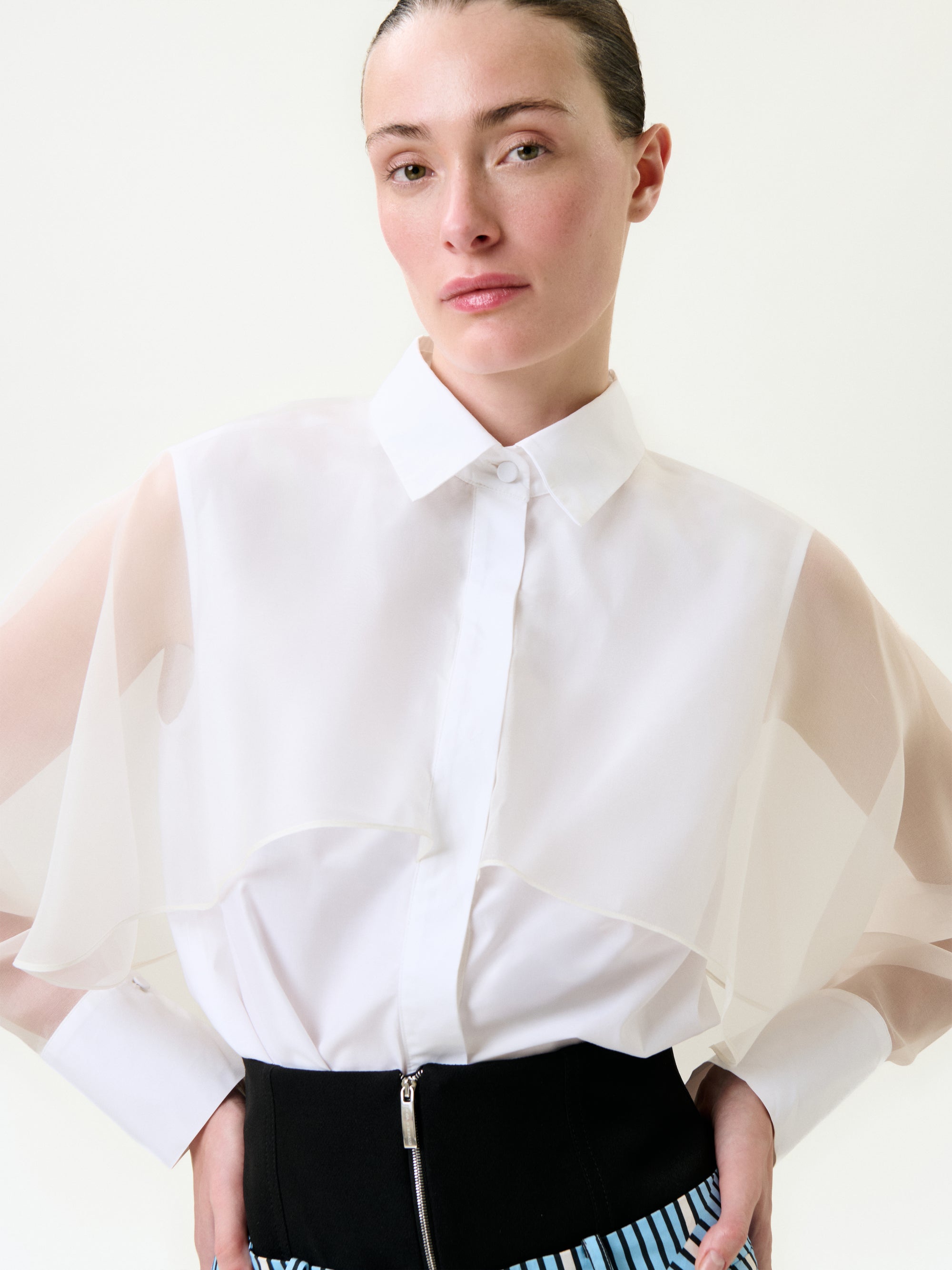 A woman with slicked-back hair wears the Nira Shirt—a sheer, layered-sleeve blouse made from 100% cotton Portuguese fabric—and a high-waisted black skirt with blue and white stripes, standing before a plain light background.