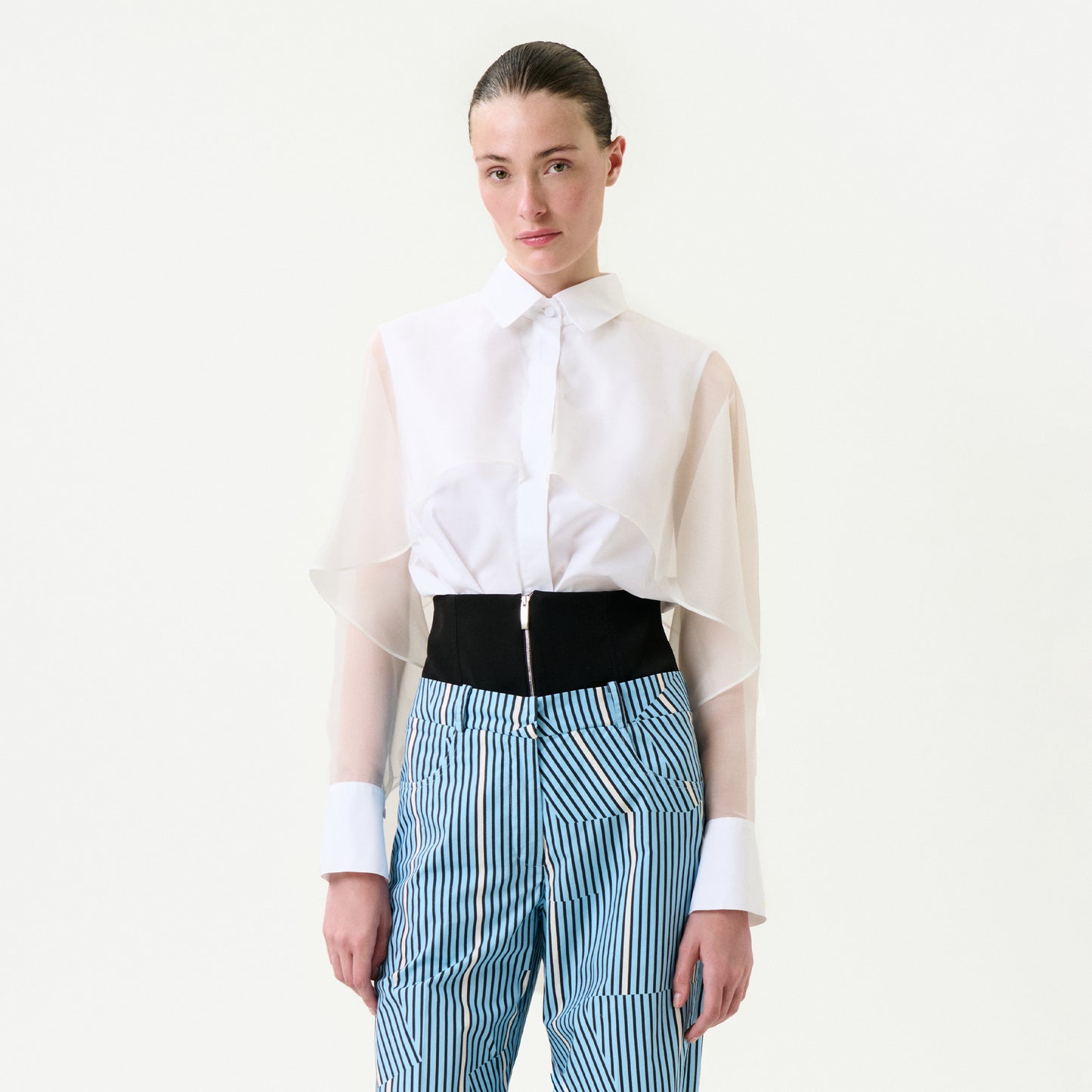 A person is wearing the Nira Shirt, a sheer white long-sleeve blouse with oversized cuffs, styled with relaxed fit blue and white vertical striped 100% cotton high-waisted pants featuring a wide black belt-like detail at the waist.