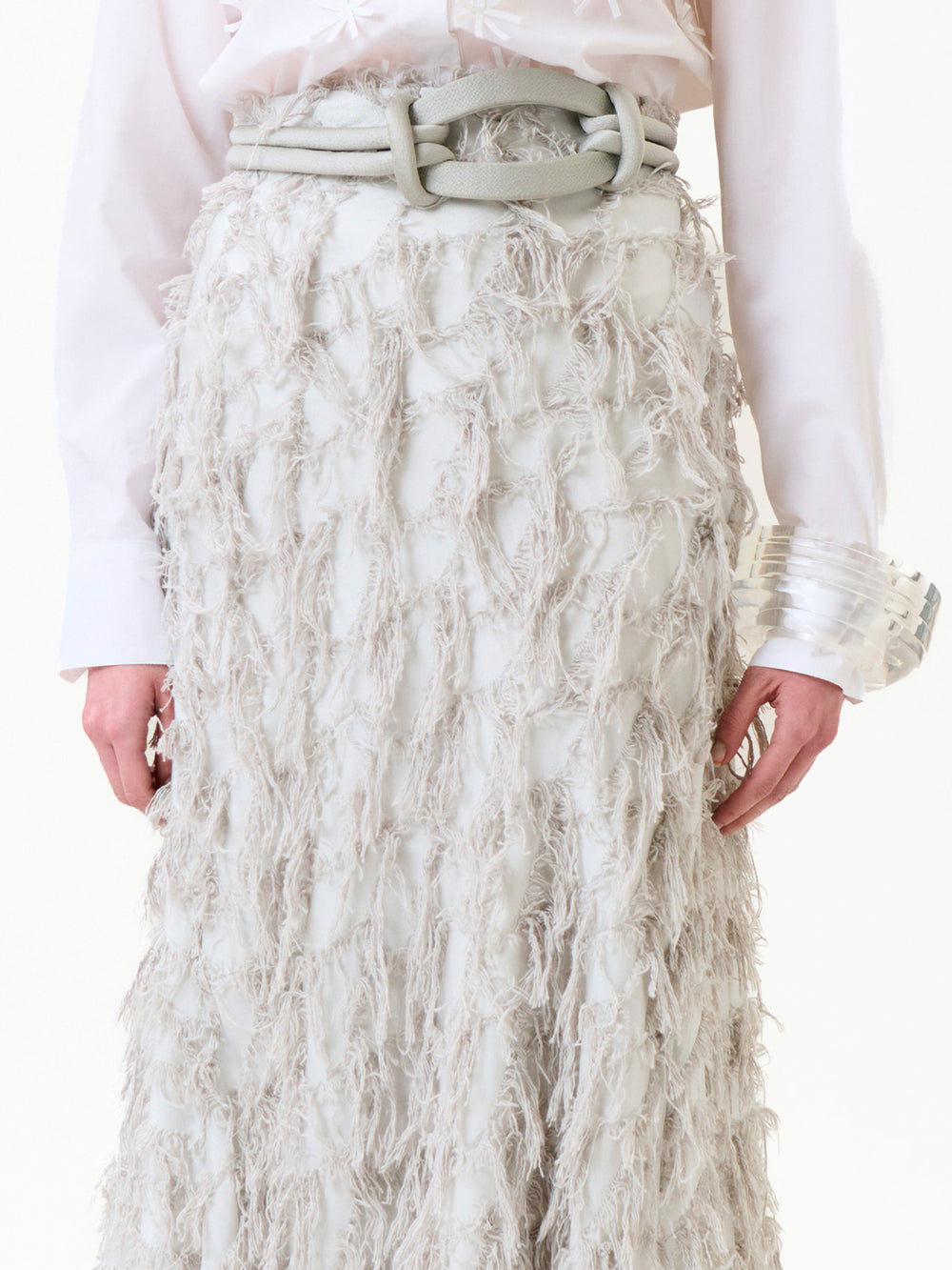 Wearing the Vania Skirt—a white textured skirt with fringe details—paired with a white belt featuring large loops, a white long-sleeve shirt, and a chunky clear bracelet on their wrist.