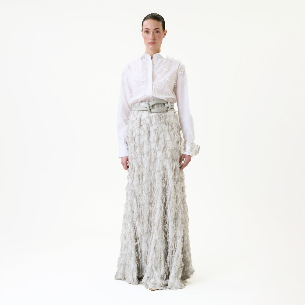 A person stands against a white background wearing the Vania Skirt—a long, textured light gray skirt—paired with a white embroidered blouse. Their dark hair is pulled back, and they are looking straight ahead with a neutral expression.