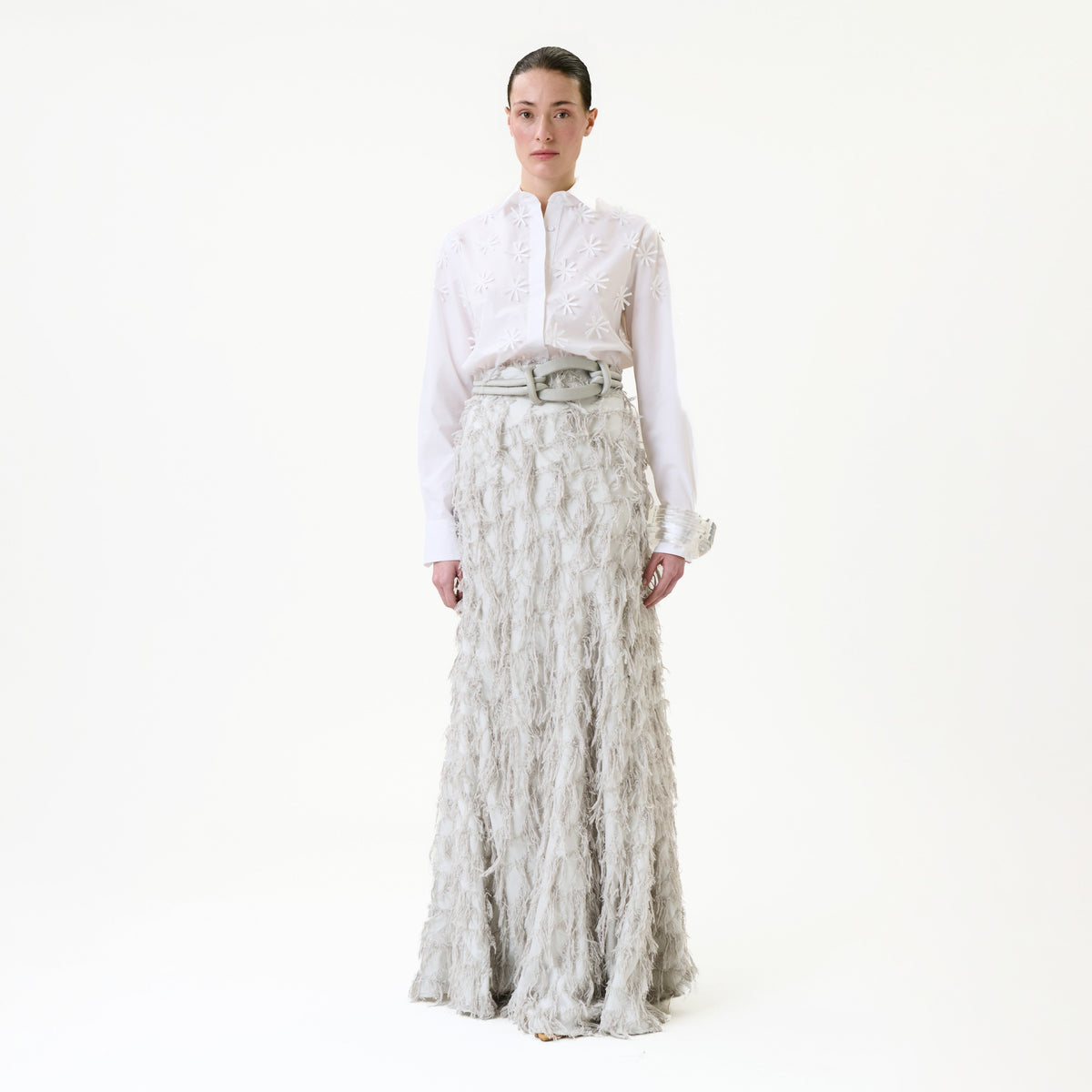 A person stands against a white background wearing the Vania Skirt—a long, textured light gray skirt—paired with a white embroidered blouse. Their dark hair is pulled back, and they are looking straight ahead with a neutral expression.