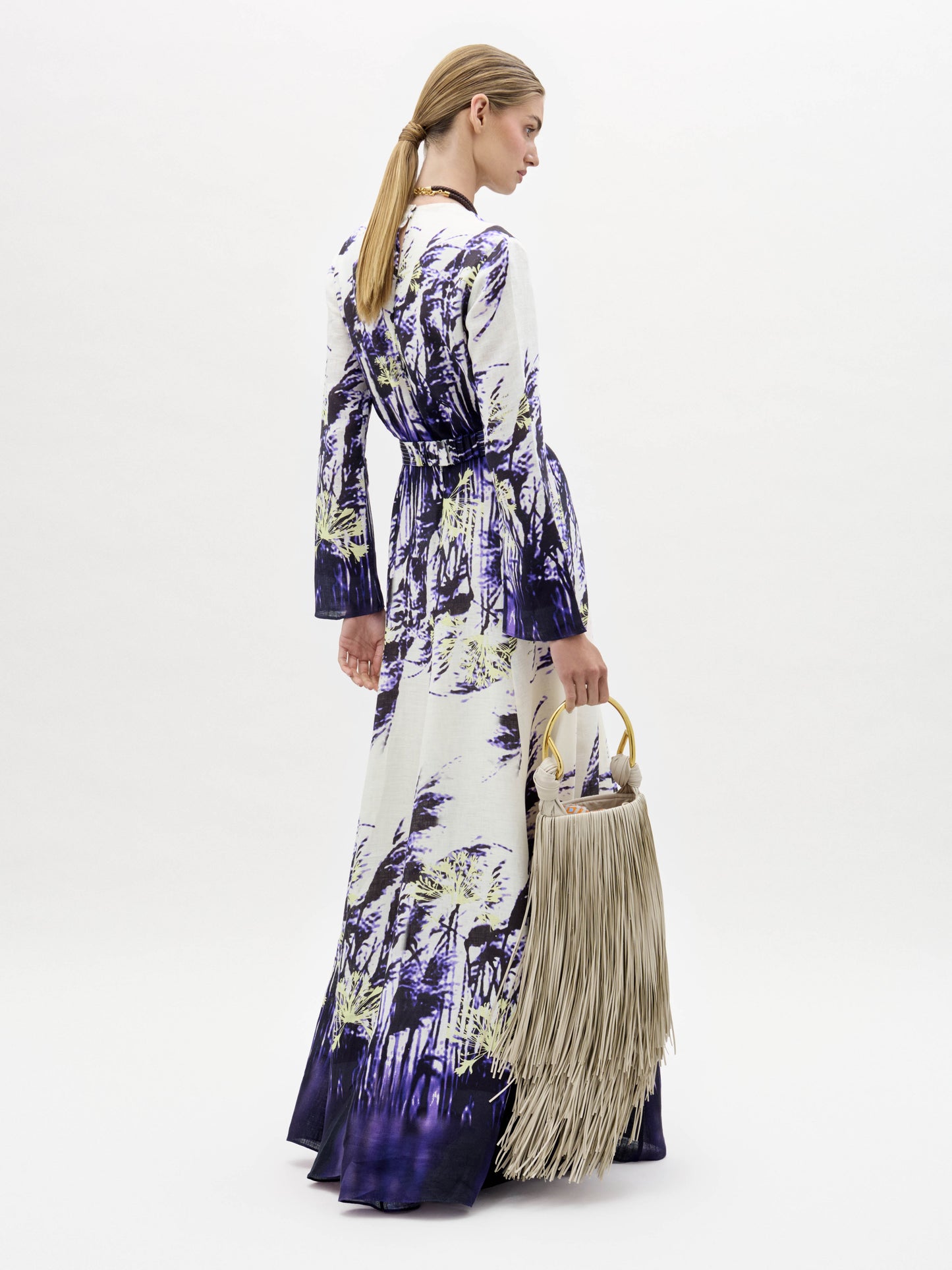 A woman in the Kady Dress, a long patterned purple and white gown, stands holding a large beige fringed handbag against a plain white background.