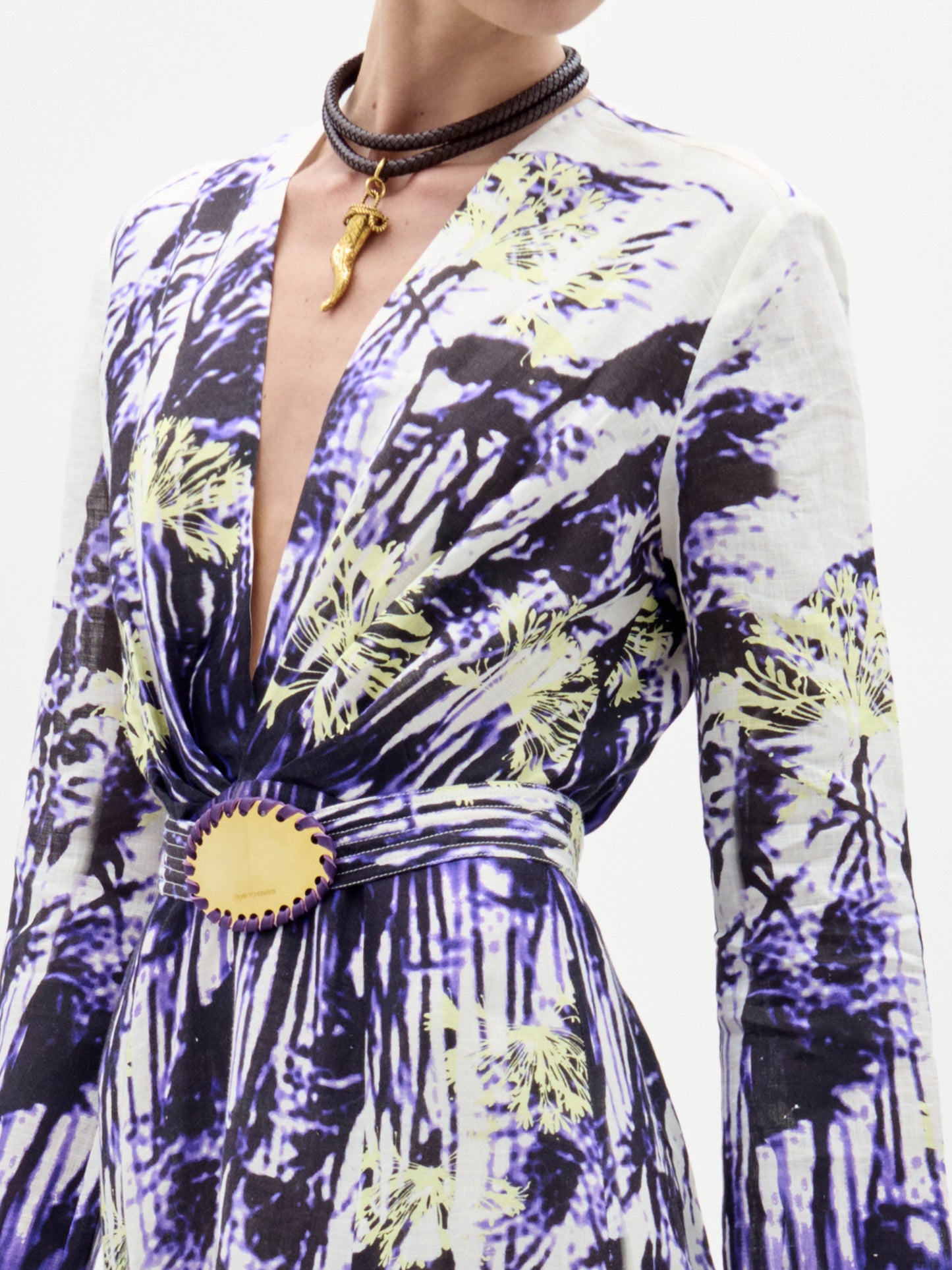A woman in the Kady Dress, a long patterned purple and white gown, stands holding a large beige fringed handbag against a plain white background.