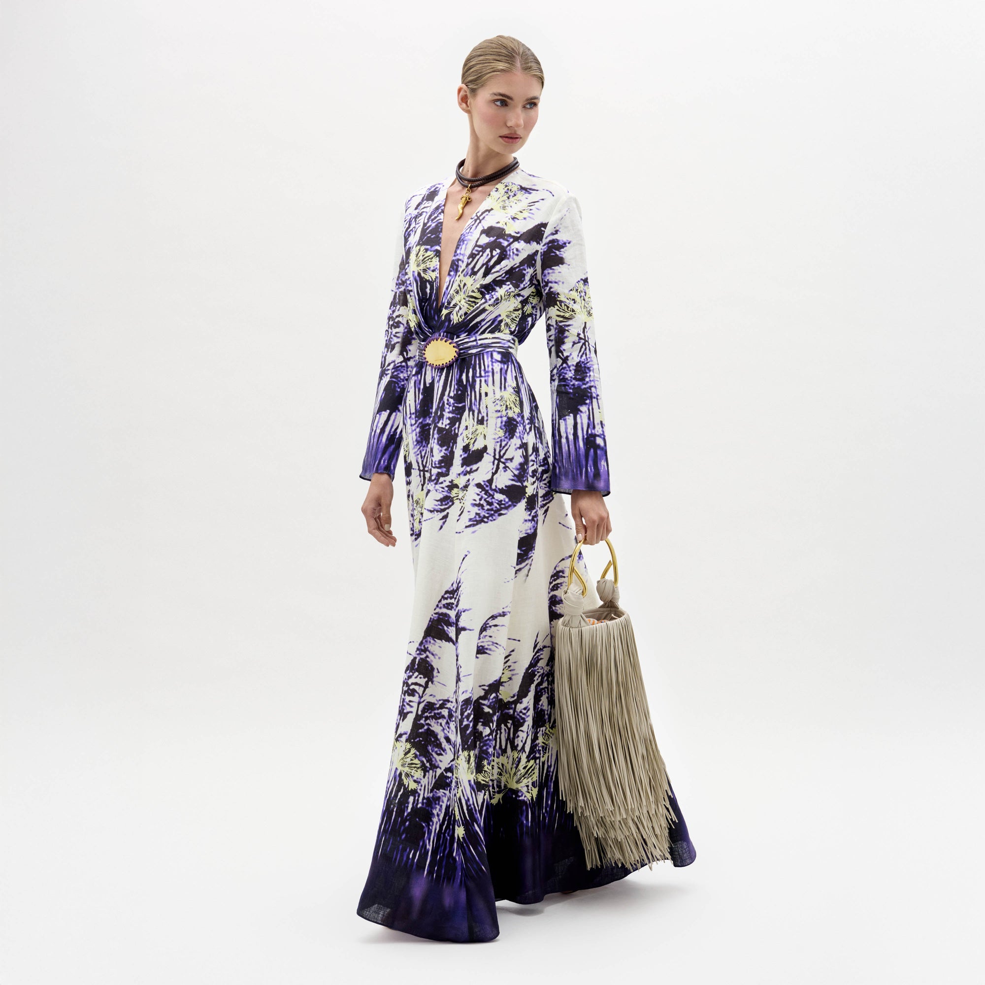 A woman in the Kady Dress, a long patterned purple and white gown, stands holding a large beige fringed handbag against a plain white background.