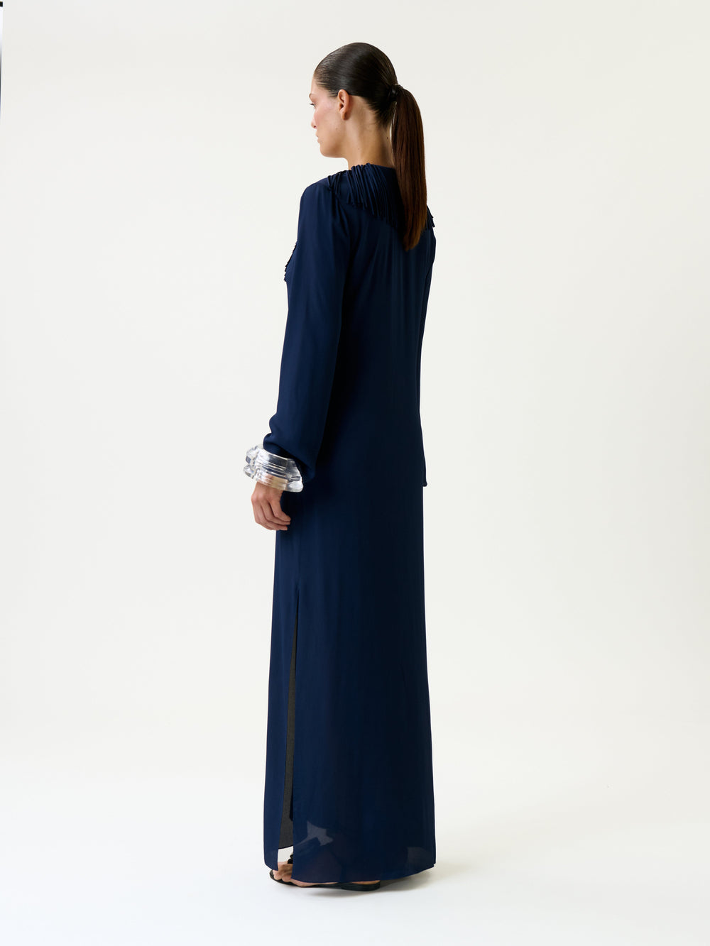 A woman stands facing away, wearing the Mercedes Dress—a long navy blue dress featuring long sleeves and a high collar. Her hair is tied in a ponytail, and she is positioned against a plain white background.