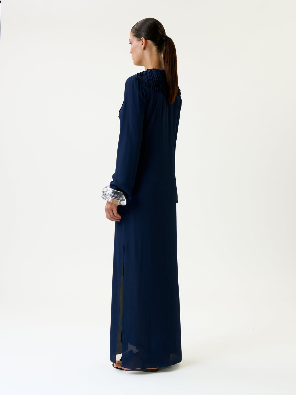 A woman stands facing away, wearing the Mercedes Dress—a long navy blue dress featuring long sleeves and a high collar. Her hair is tied in a ponytail, and she is positioned against a plain white background.