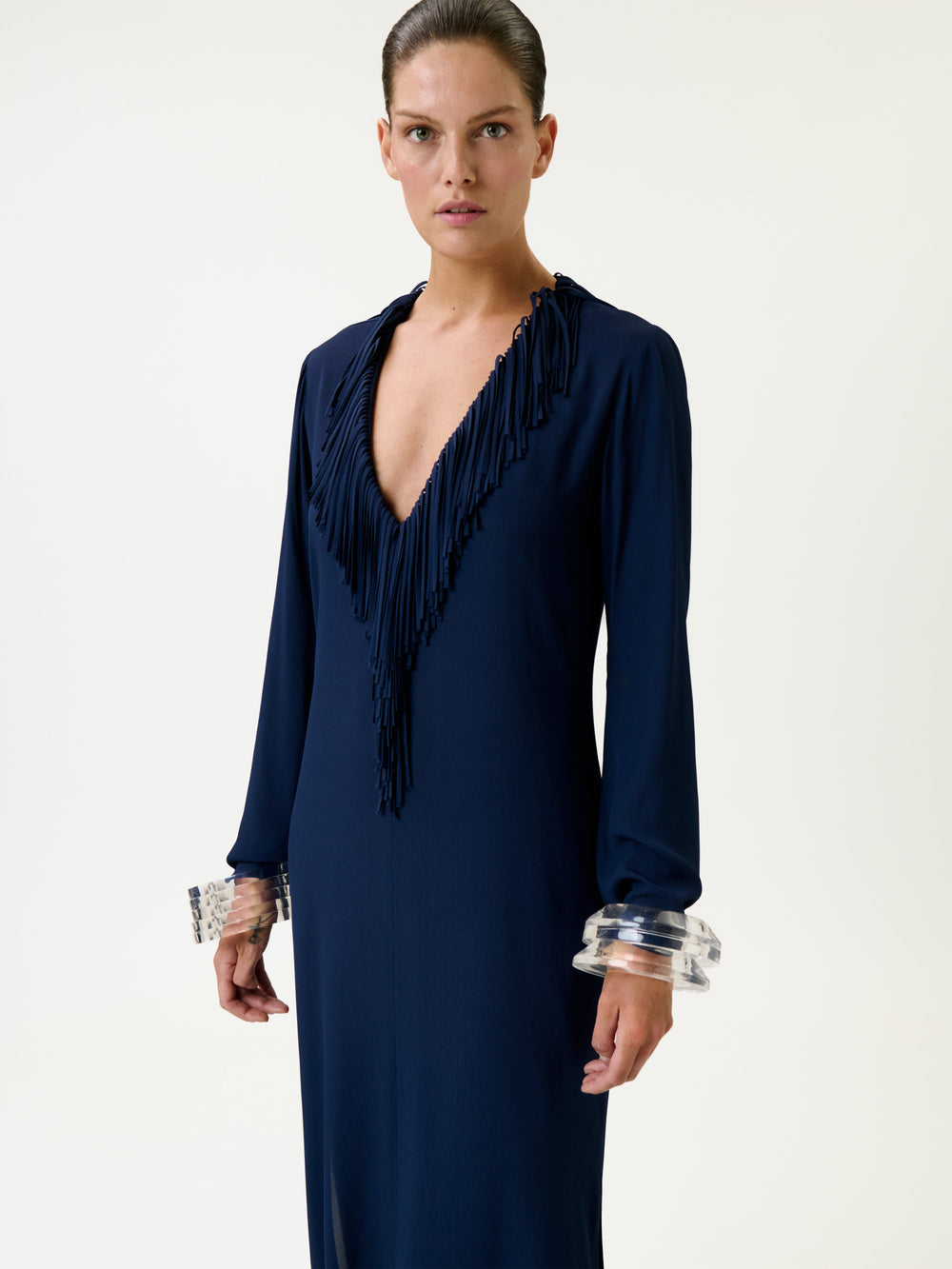 A person wearing the Mercedes Dress in navy blue, featuring a deep V neckline and fringe detail, stands against a plain background. They have slicked-back hair and clear, chunky bracelets on each wrist.