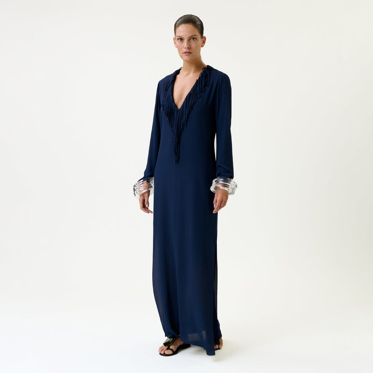 A person stands against a white background wearing the Mercedes Dress, a long, deep navy blue style featuring fringe details at the neckline, paired with clear chunky bracelets and black sandals.