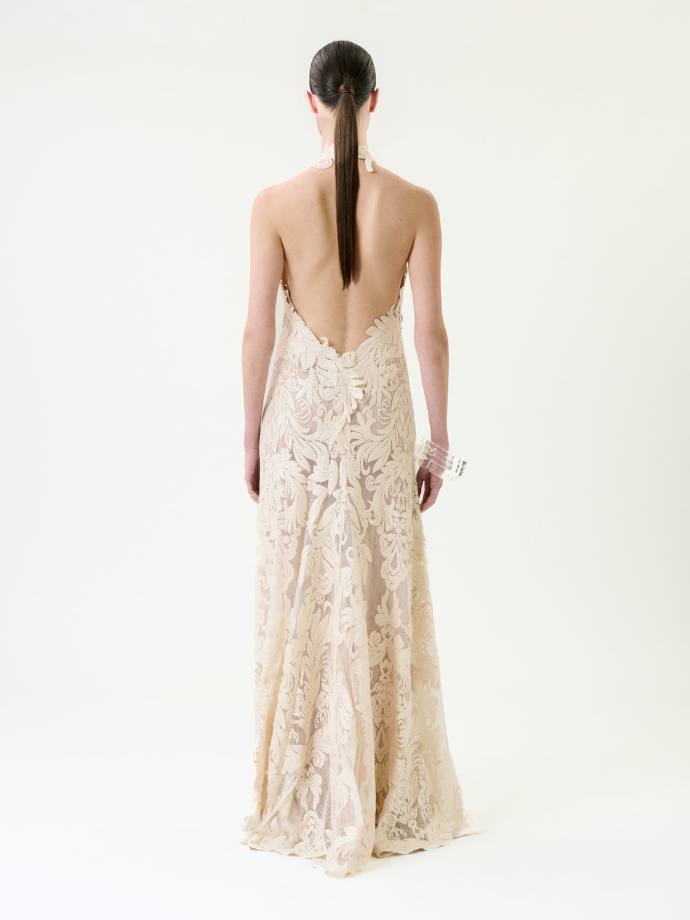 A person with long, straight brown hair in a low ponytail wears the Marcia Dress—a backless, floor-length cream lace gown with a floral pattern—standing against a plain white background.