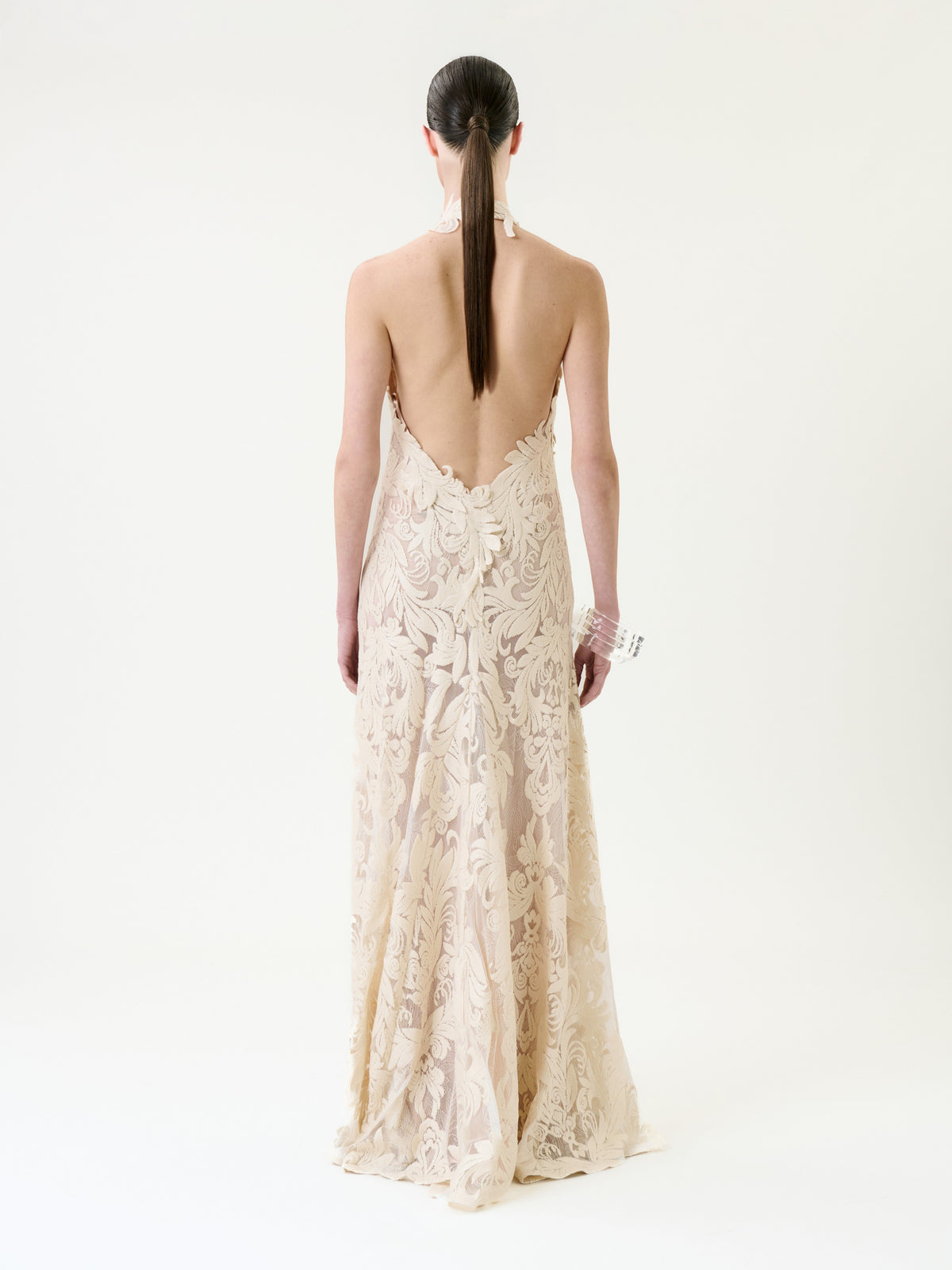 A person with long, straight brown hair in a low ponytail wears the Marcia Dress—a backless, floor-length cream lace gown with a floral pattern—standing against a plain white background.