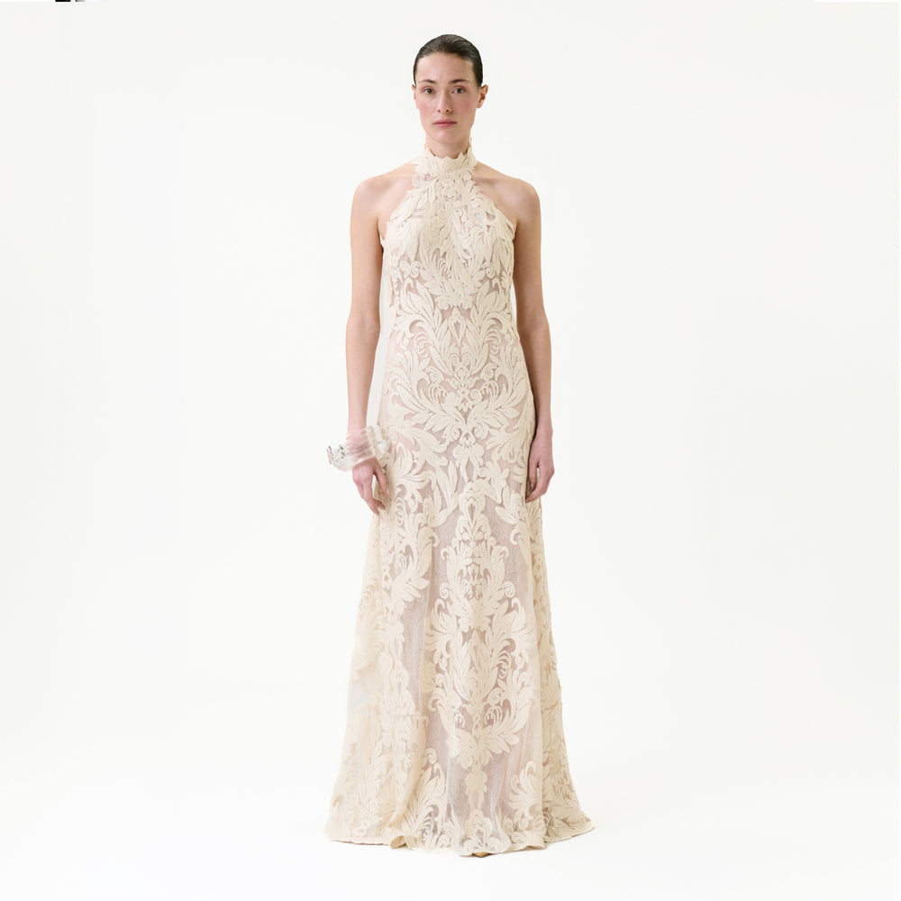 A woman faces forward wearing the Marcia Dress, a sleeveless, floor-length cream gown adorned with intricate lace patterns. The background is plain and white.