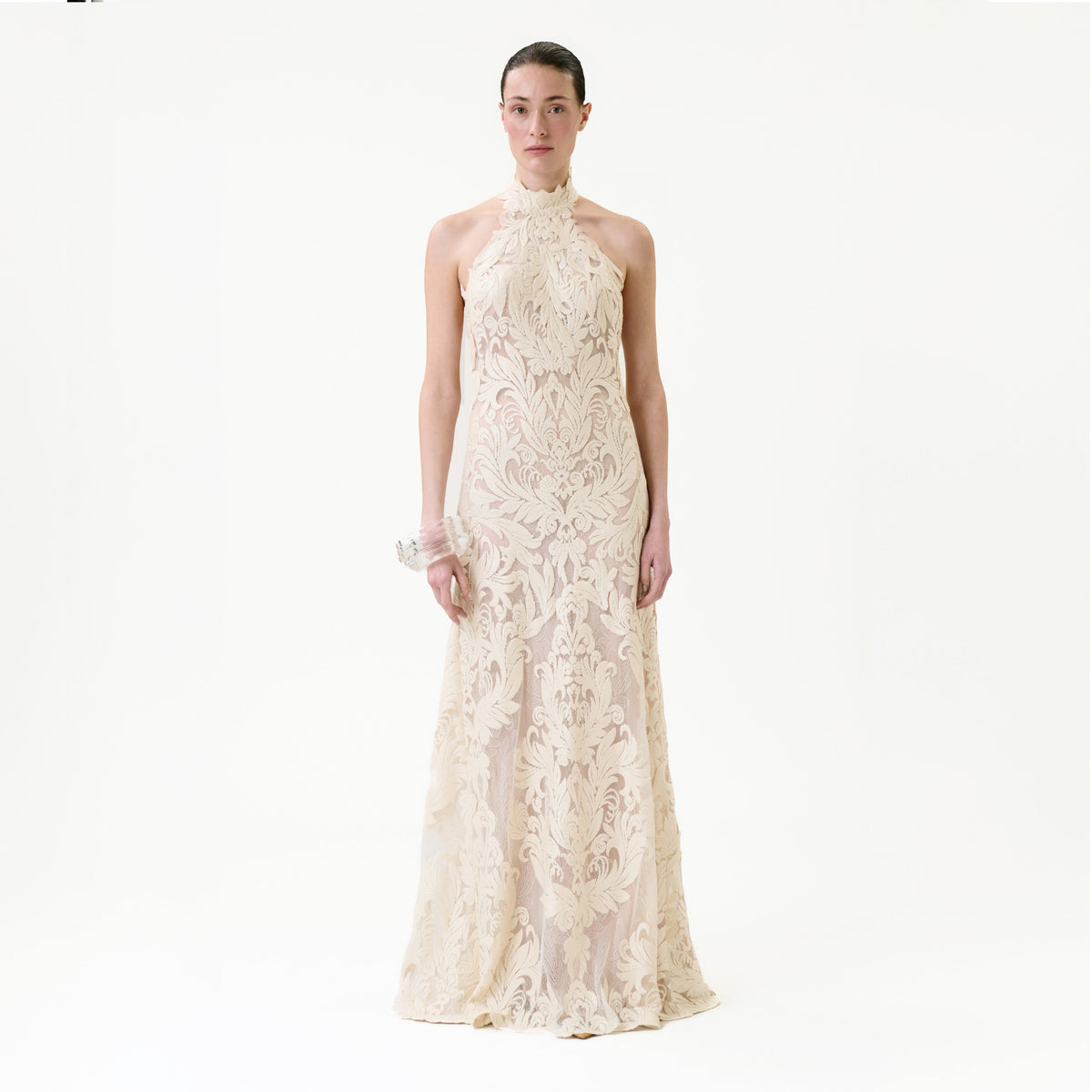 A woman faces forward wearing the Marcia Dress, a sleeveless, floor-length cream gown adorned with intricate lace patterns. The background is plain and white.