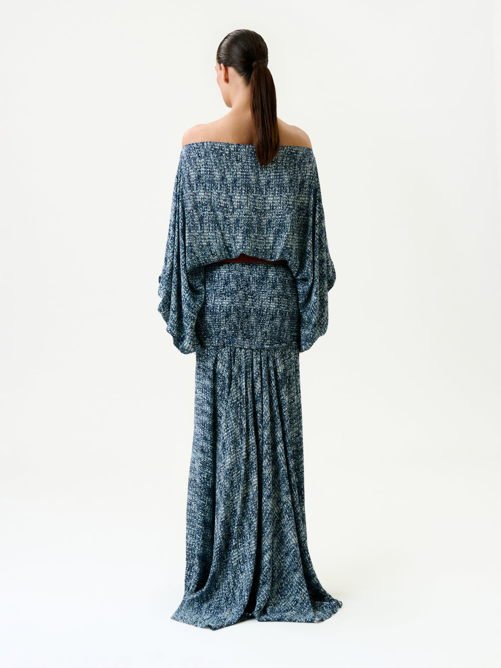 A person with long dark hair in a low ponytail faces away, wearing the Nelly Dress—an off-shoulder, blue-gray textured maxi dress with loose sleeves and a flowing skirt—made in Colombia, against a plain white background.