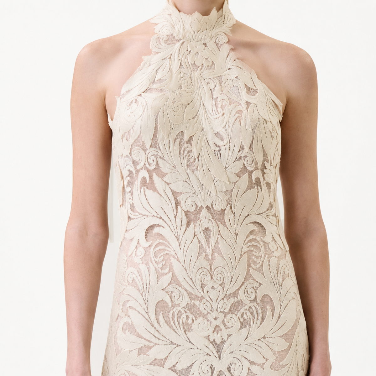 A person wearing the Marcia Dress, a sleeveless high-neck design featuring intricate cream lace floral patterns, standing against a plain light background.