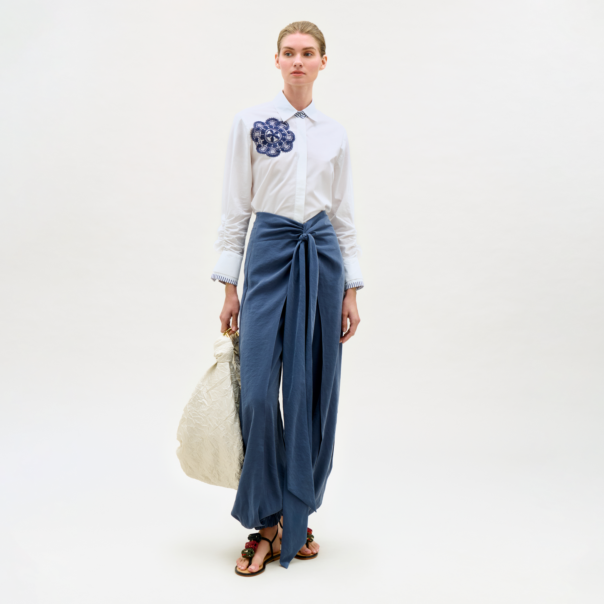 Woman wearing a white shirt with a floral design and blue pants, holding a beige bag on a white background