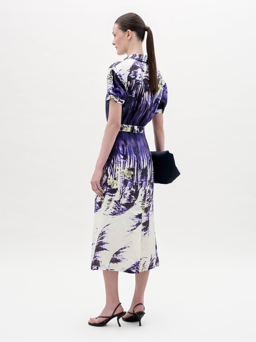 A woman wears the Lee Dress, a short-sleeve purple and white patterned dress with a belt, paired with a black clutch and black sandals, standing against a plain white background.