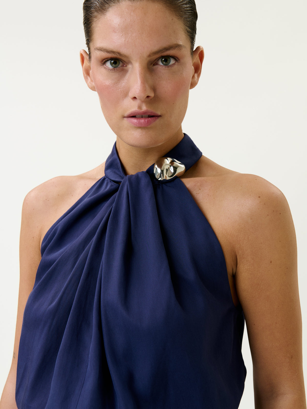 A person with slicked-back hair models the Shela Blouse, a navy halter-top featuring a silver decorative clasp at the neck, standing against a plain light background.