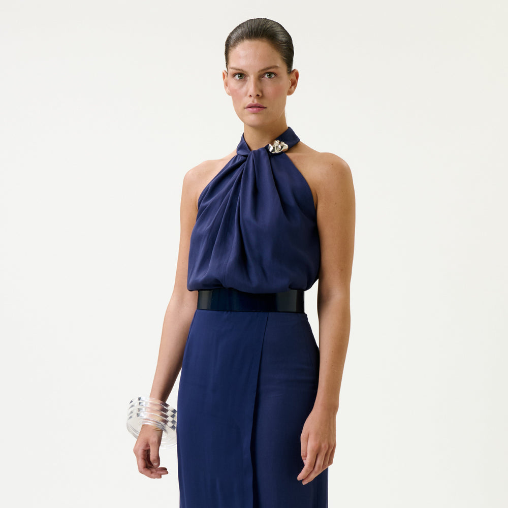 Wearing the Shela Blouse, a woman stands against a plain background, styled with a matching skirt, wide black belt, and clear bangles.