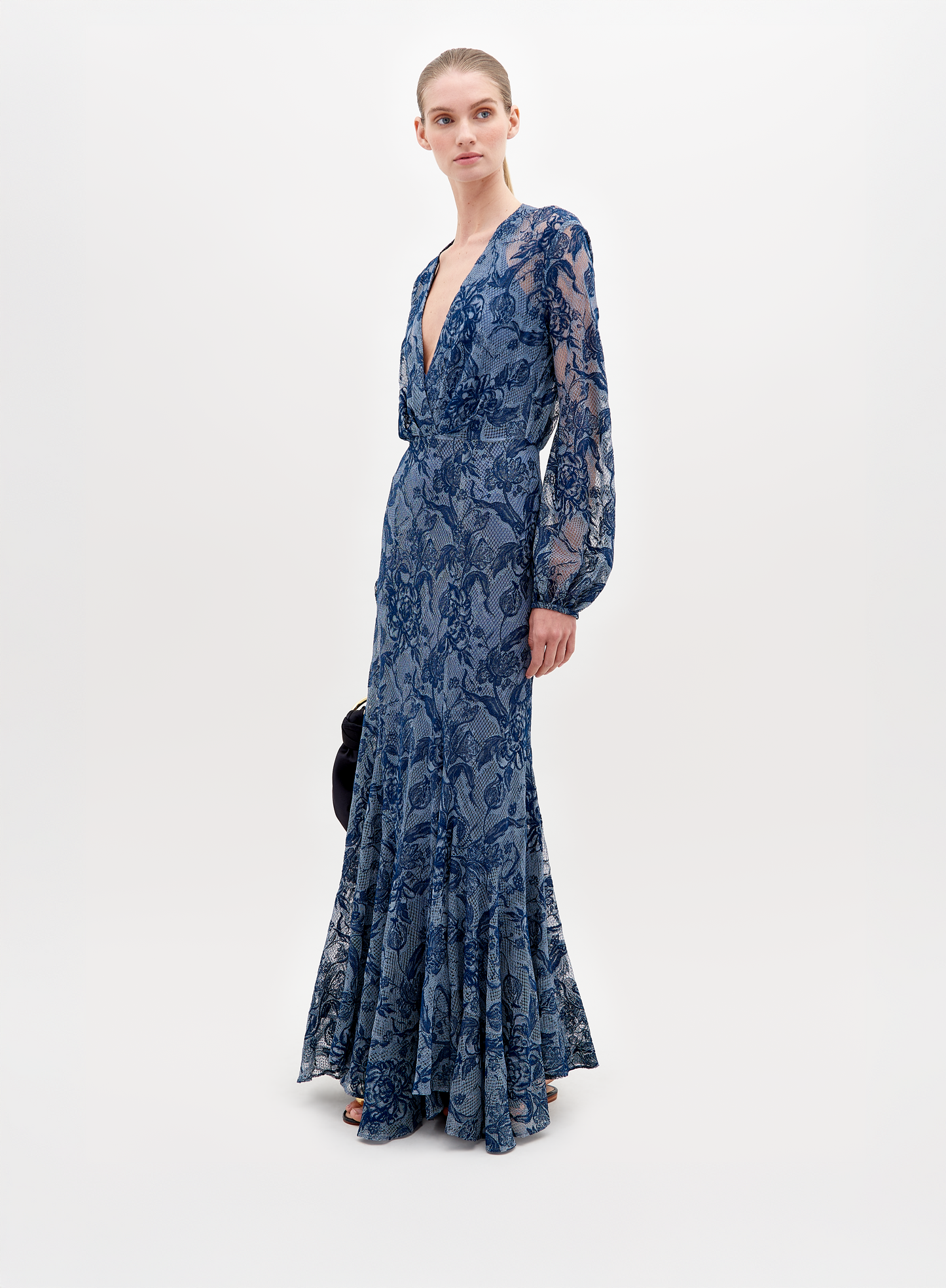 A person in a long, blue lace dress with a deep V-neckline and long sleeves stands against a plain white background, holding a black clutch—the Robin Dress Indigo Lace, available for pre-order, set to ship by November 15th, 2024.