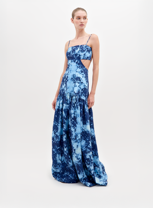 A woman stands wearing a long, blue, tie-dye sleeveless dress with thin straps and side cutouts, posing against a plain white background. The Shannon Dress Indigo is available for pre-order now and will ship by November 15th, 2024.