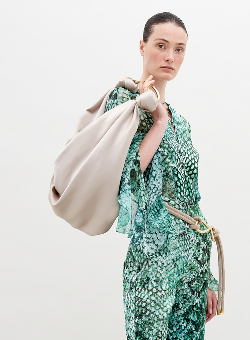 A person with slicked-back hair wears Thea Blouse Emerald Animal Print and holds a large beige bag over their shoulder, perfect for those looking to pre-order now. The background is plain white, highlighting the stylish ensemble expected to ship by November 15th, 2024.