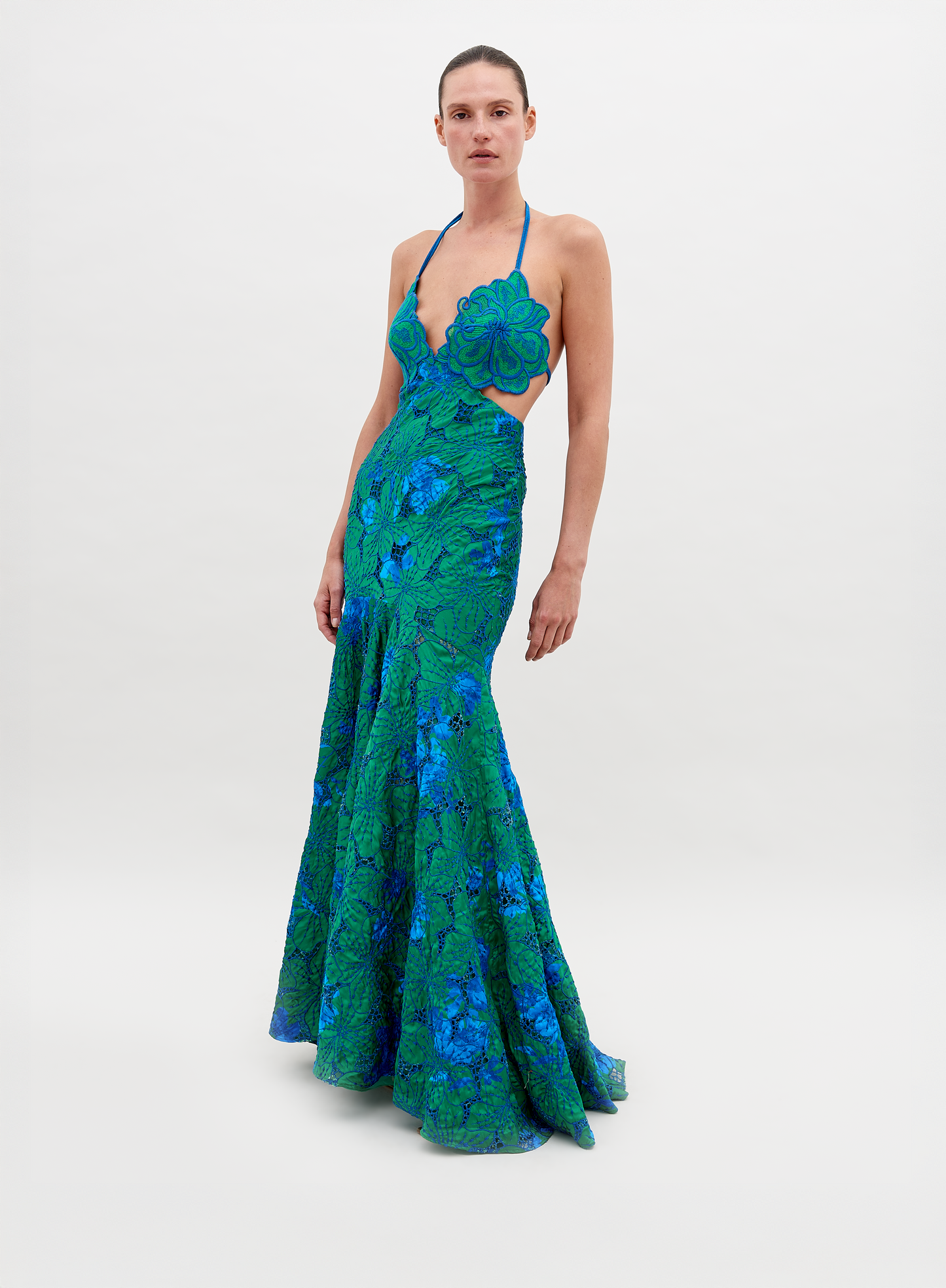 A person stands wearing a vibrant green and blue floral-patterned dress with an open back and a halter neck, posing against a plain white background. The Cara Dress Green Navy Embroidery is available for pre-order now, with a ship by date of November 15th, 2024.