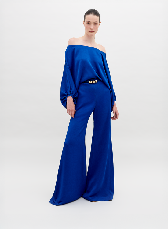 A person stands against a plain background wearing an off-the-shoulder blue top with wide sleeves and matching wide-leg pants, accessorized with a belt that has two large gold embellishments. Pre-order the Theresa Jumpsuit Royal Blue now to ensure you receive your outfit by the ship by date of November 15th, 2024.