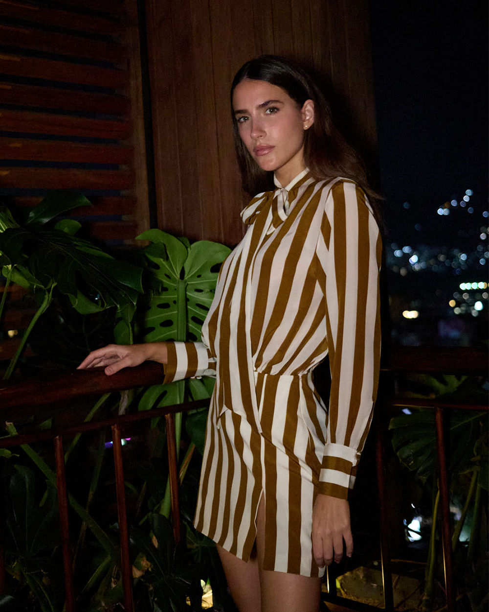 A woman wearing a brown and cream vertically striped long-sleeve shirt dress, posing on a balcony at night with plants and city lights in the background.