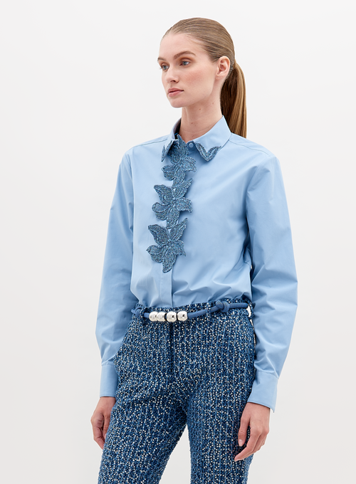 A woman with straight hair in a ponytail wears a Naroa Shirt Celestial Blue with ruffled detailing on the front and textured blue high-waisted pants, accessorized with a white beaded belt. Available for pre-order now, with an expected ship date in November 2024.