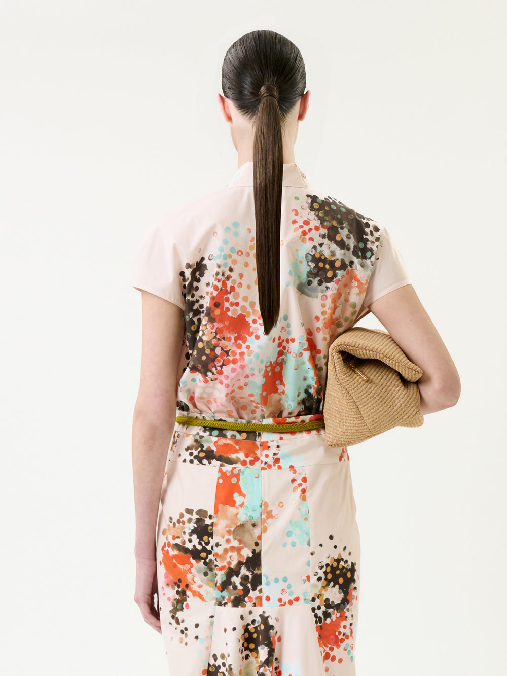A woman with a sleek ponytail, seen from behind, wears the Maribel Shirt—a short-sleeve top featuring a colorful abstract dot pattern—and holds a tan textured clutch.
